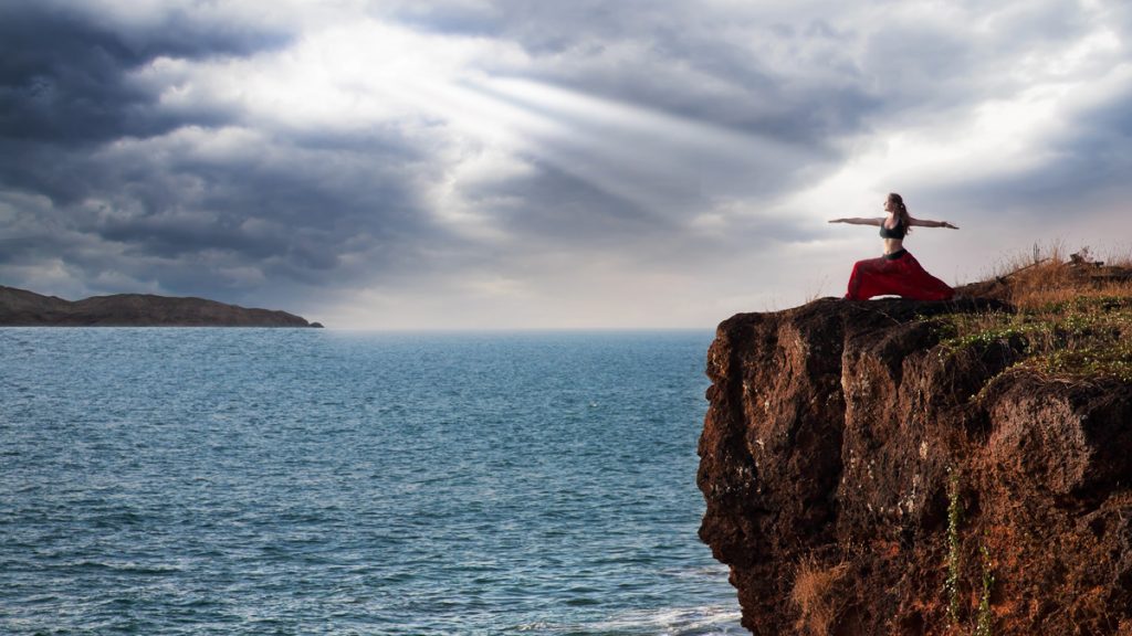 Beautiful woman doing virabhadrasana warrior yoga pose on the cliff near the ocean with dramatic sky at background in India