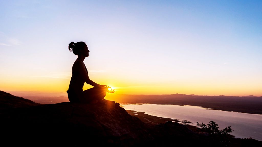 A woman with yoga posture on the mountain at sunset; Silhouette of gorgeous young woman practicing Lotus yoga outdoor.