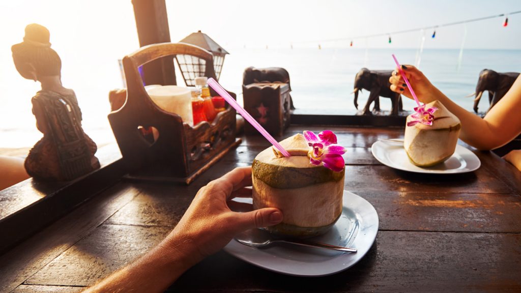 Couple drinks sweet fresh coconuts decorated with flower at restaurant on the beach at sunrise